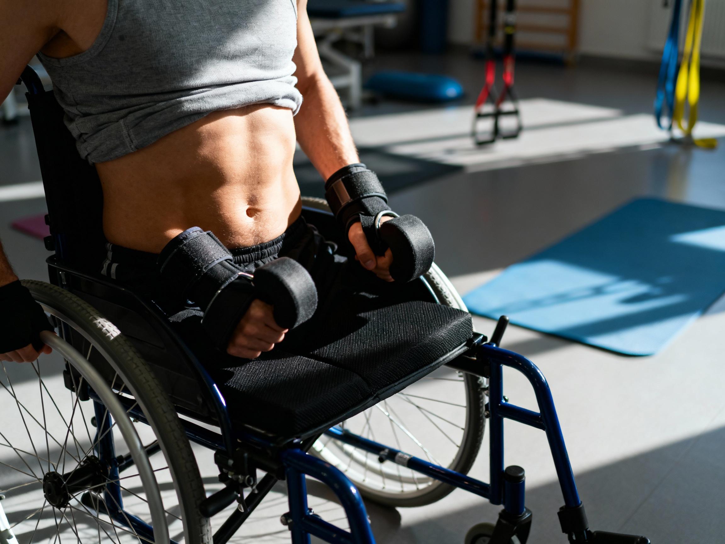 Close-up of a person’s torso in a wheelchair, showcasing core muscle tension while lifting weighted cuffs, with dynamic lighting emphasizing abdominal engagement. The background is a blurred gym environment with therapy mats and resistance bands. Keywords: core stability, wheelchair exercise, adaptive physiotherapy.